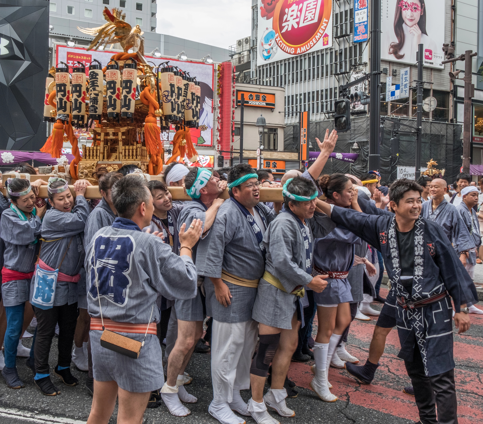 A group of people in traditional Japanese festival clothing carry a portable shrine (mikoshi) during a lively autumn street festival in an urban area with many signs and buildings visible in the background.