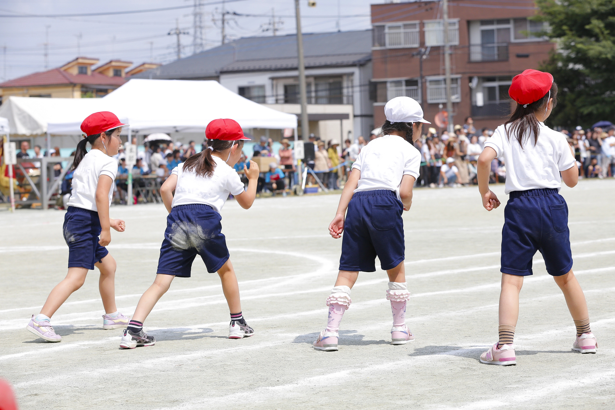 Four children in Japanese PE uniform of white shirts, navy shorts, and red or white caps line up on a sports field, preparing for a race, with spectators and tents in the background—a lively scene typical of the sports festivals (undokai) held every October in Japan.