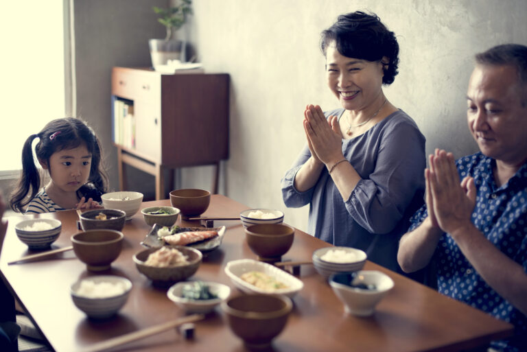 Three people sit at a dining table with various dishes, their hands together in a gesture of gratitude before a meal, in a Japanese home