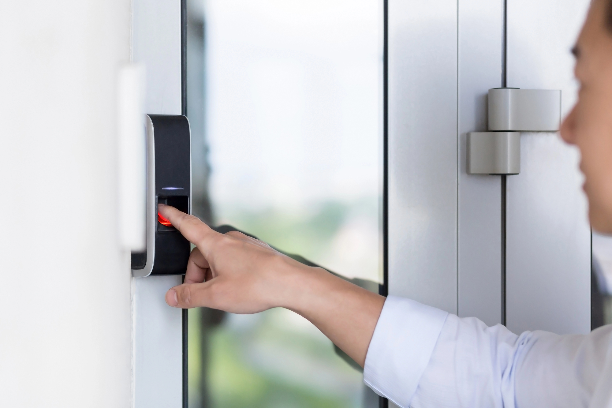 Person pressing a fingerprint scanner on a wall-mounted access control device next to a glass door, much like following proper phrases for visiting a Japanese home—demonstrating both respect and security upon entering.