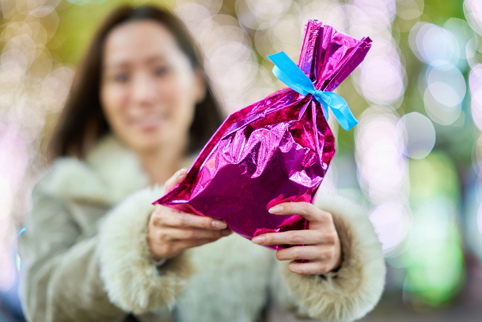 A person in a fur-trimmed coat holds out a shiny pink gift bag wrapped with a blue ribbon—an elegant gesture reminiscent of the careful phrases for visiting a Japanese home. The background is softly blurred with lights, adding warmth and subtle hospitality.
