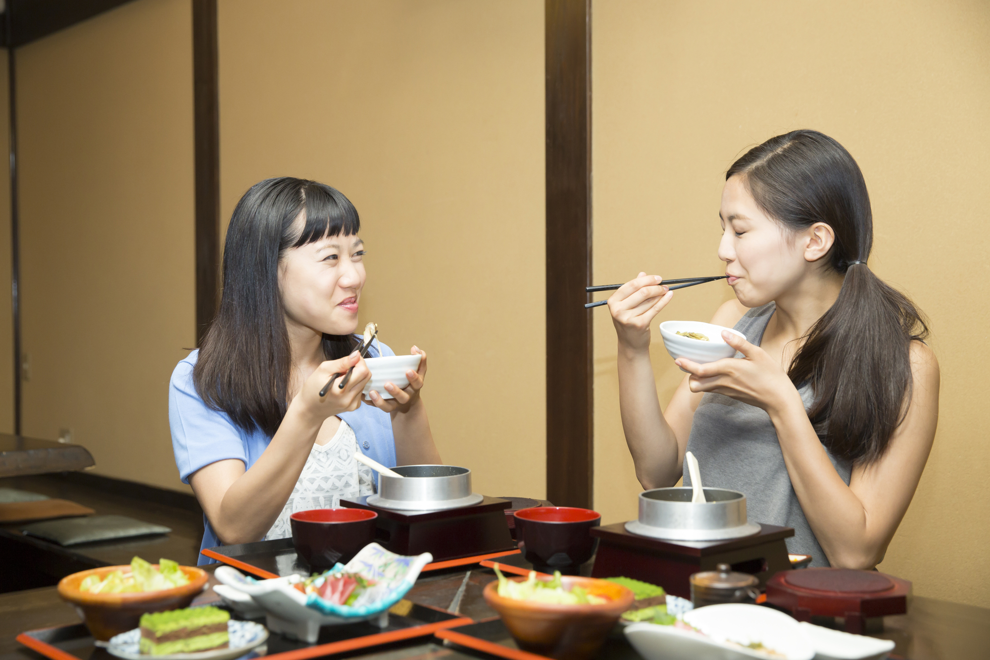 Two young women sit at a table in a restaurant, eating with chopsticks and bowls, with various dishes in front of them. As they enjoy their meal, they practice phrases for visiting a Japanese home, sharing language tips and cultural insights in between bites.