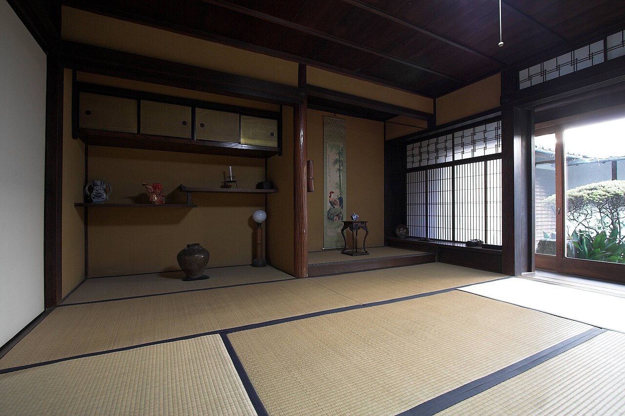 Traditional Japanese tatami room with sliding shoji doors, wooden ceiling, built-in shelves, scroll, and decorative objects, with sunlight entering from a garden outside.