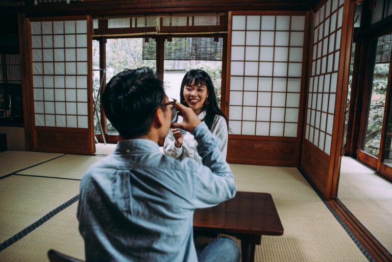 Two people sit at a low wooden table in a traditional Japanese-style room with shoji screens and tatami mats, smiling and talking.