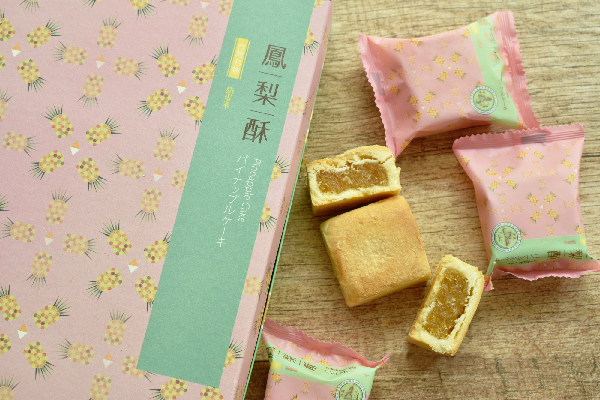 A box and three individually wrapped pineapple cakes, with two cakes cut open showing their golden filling, displayed on a wooden surface.