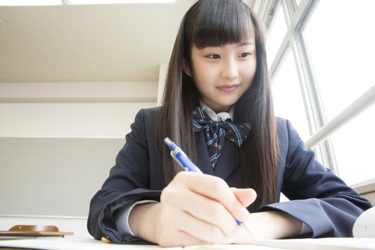 A Japanese student in a school uniform writing with a pen at a desk.